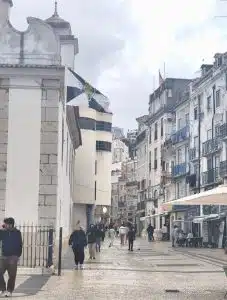Cena de rua na Rua da Mouraria, em Lisboa, com edifícios históricos, peões e passeio molhado após a chuva.
